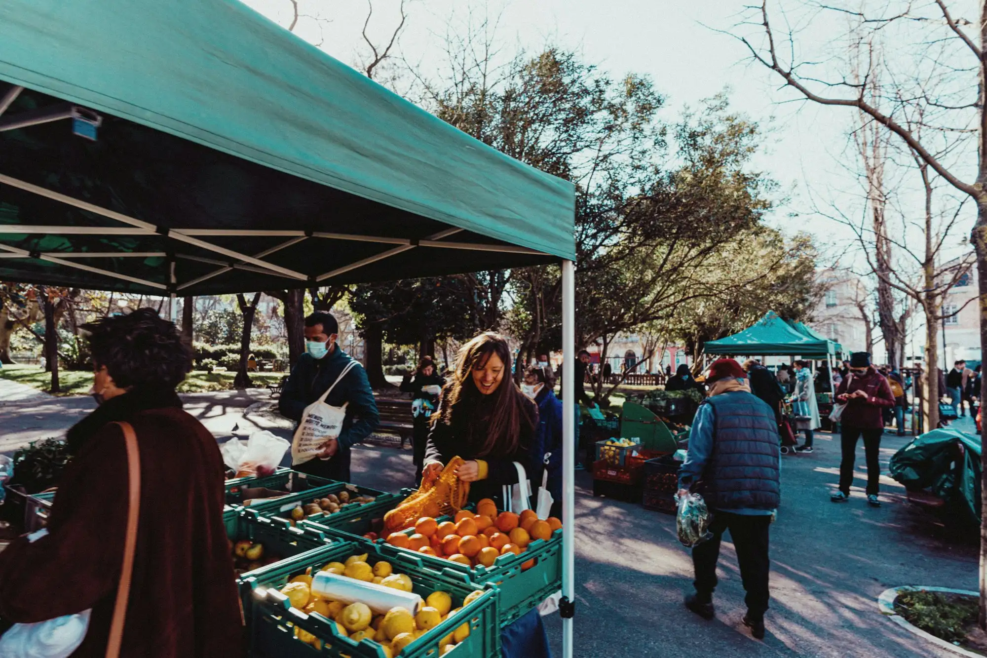 Shoppers browsing fruit and vegetables under a green canopy at an outdoor farmers market