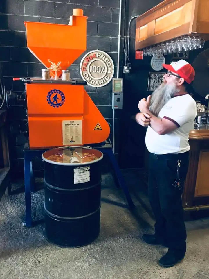 The owner of Boldt Alchemy Distillery looks at a machine that makes Boldt spirits, in front of a sign reading “Farm Fresh 100% Organic” and a wooden bar with glasses