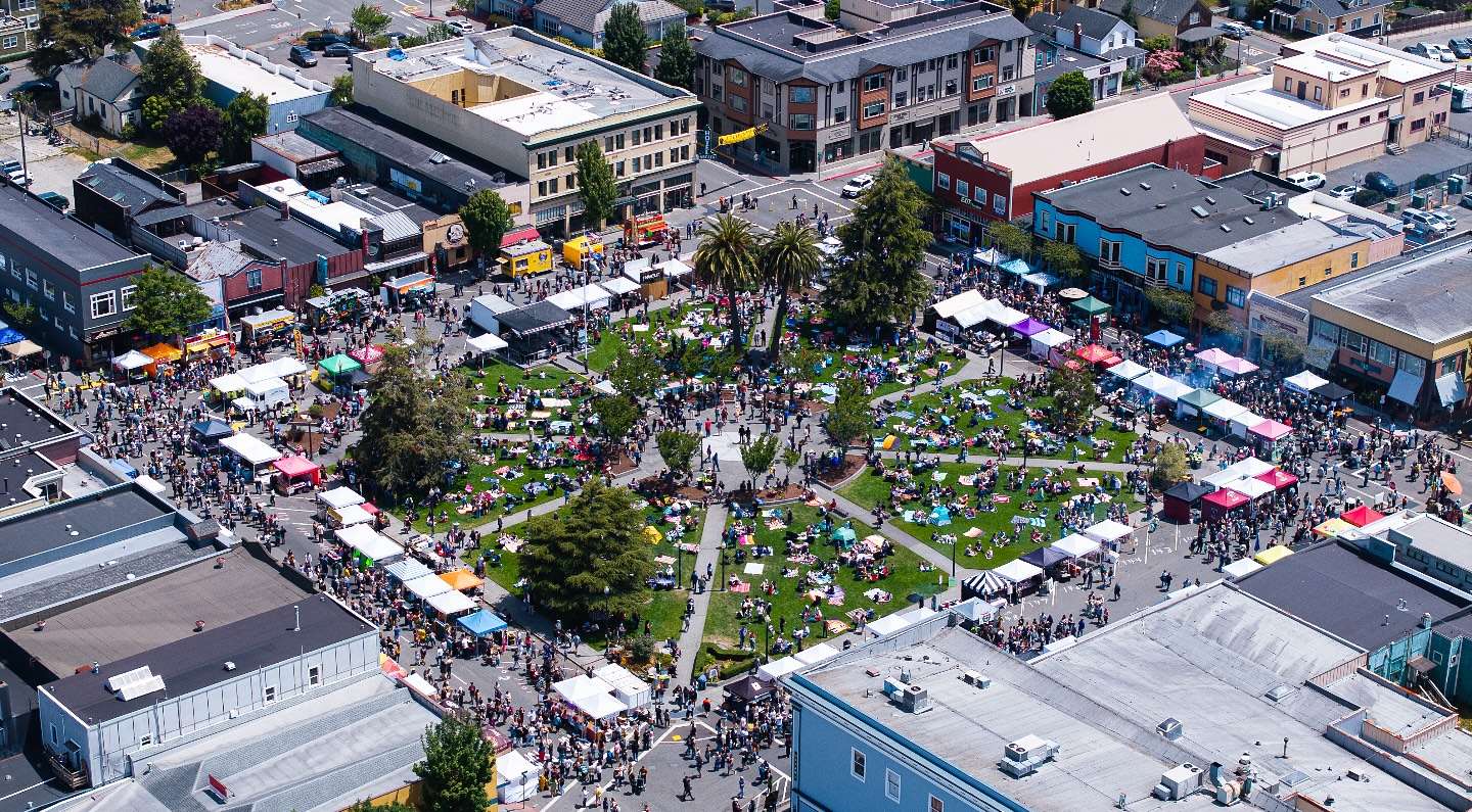 Aerial view of a huge crowd filling the Arcata Plaza, with palm trees, vendor tents, and an American flag, during the Oyster Festival