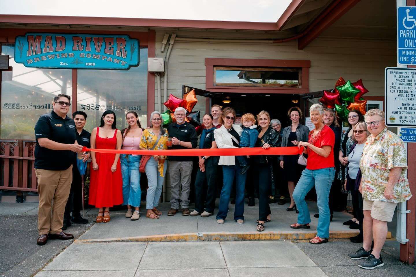 Chamber members and neighbors hold a red ribbon outside Mad River Brewing Company, decorated with balloons