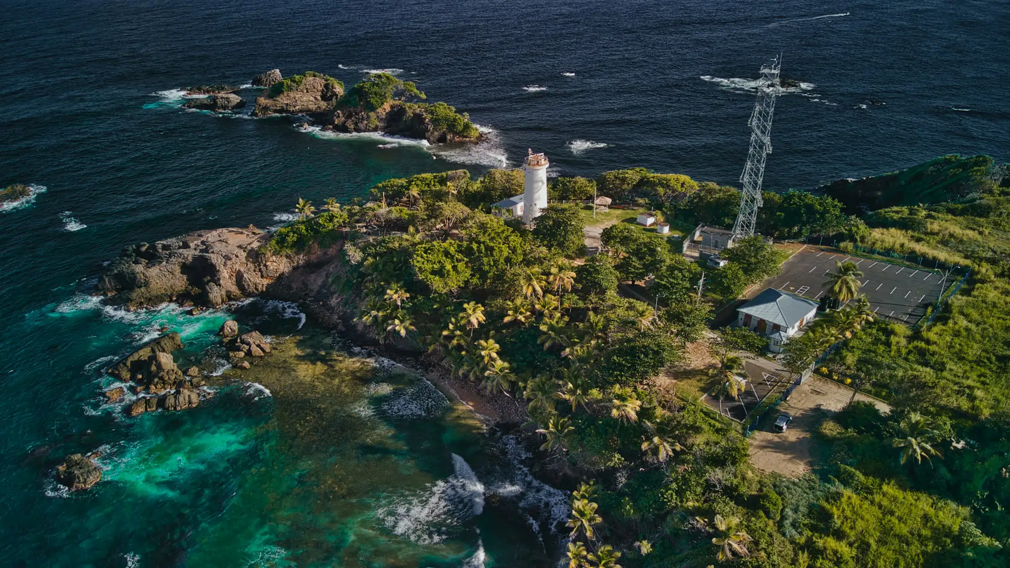 Aerial view of a small white lighthouse and a tall cross on a forested headland above turquoise water