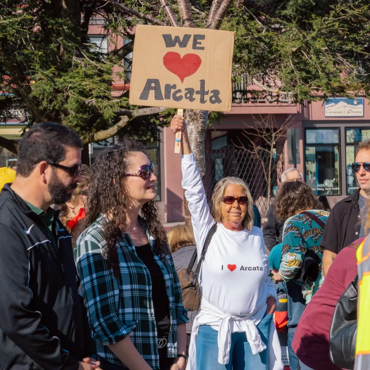 A smiling woman at a downtown gathering holds a cardboard sign reading “We love Arcata”